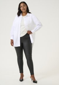 Weißes Hemd mit Knopfleiste, über einem hellen Pullover getragen, kombiniert mit schwarzen Leggings aus Kunstleder und offenen schwarzen High Heels. Einfaches Design, figurbetonte Form.