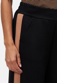 Des pantalons de sport noirs présentent une bande beige sur le côté, une texture douce, une taille élastique et des poches latérales. Gros plan sur une main sur le pantalon.
