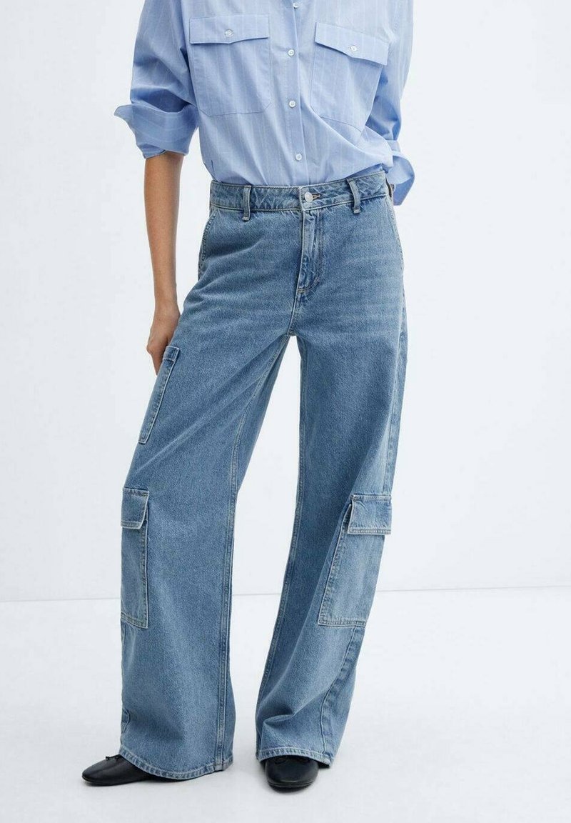 Personne portant une chemise bleu clair boutonnée et un pantalon cargo en denim bleu à jambes larges avec poches plaquées et des chaussures noires, debout devant un fond uni.