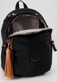 Zaino nero con scomparto principale aperto, zip nella tasca frontale, hardware rosa antico e grande portachiavi a nappina beige attaccato alla zip.