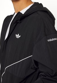 Veste noire à capuche en tissu léger, avec des accents blancs et le logo Adidas. Fermeture éclair et détails de capuche réglable.