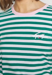 Zielona i biała koszulka w paski z różowym kołnierzykiem. Posiada wyszywane logo "Tommy" z małym akcentem flagi. Wykonana z miękkiego materiału bawełnianego.