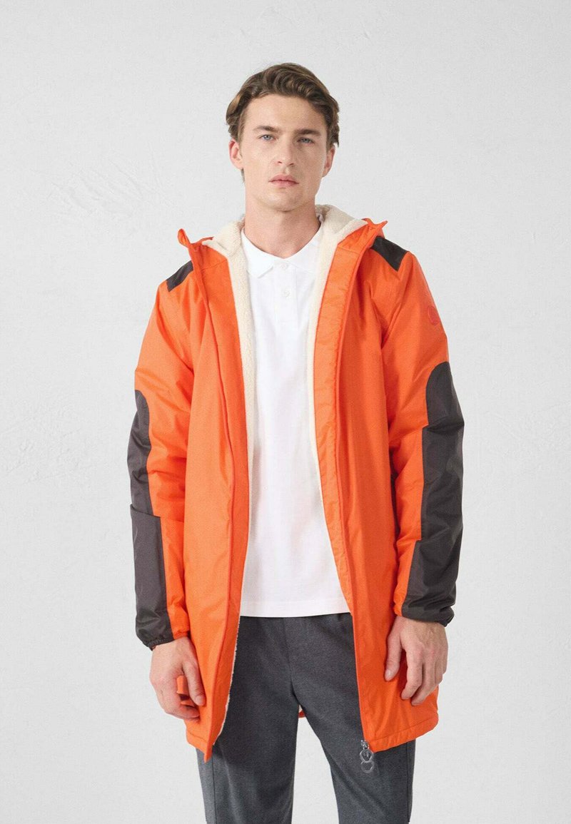 Jonge man die een open feloranje jas draagt met zwarte elleboogstukken, een wit poloshirt en donkergrijze broek, staande tegen een effen achtergrond.