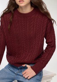 Maglione a trecce color bordeaux con scollatura rotonda e polsini a coste, abbinato a jeans in denim blu chiaro. Dettaglio della superficie testurizzata visibile.