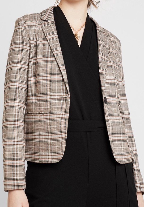 Femme portant un blazer à carreaux beige sur une robe portefeuille noire à col en V, accessoirisée d'un collier en chaîne dorée et de boucles d'oreilles géométriques.