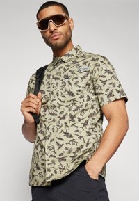 Korte mouwen button-up shirt in lichtgroen met donkerbruine patronen geïnspireerd op de natuur. Bevat een borstzakje en een merklabel.