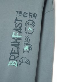 Sudadera gris con el texto "ES HORA DEL DESAYUNO", adornada con íconos de un reloj, una taza humeante y un croissant en acentos negro y verde menta.