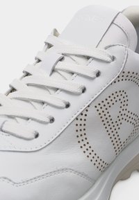 Κοντινή λήψη ενός λευκού δερμάτινου αθλητικού παπουτσιού με λευκά κορδόνια και διάτρητη λεπτομέρεια στο πλάι που σχηματίζει ένα σχέδιο ή λογότυπο.