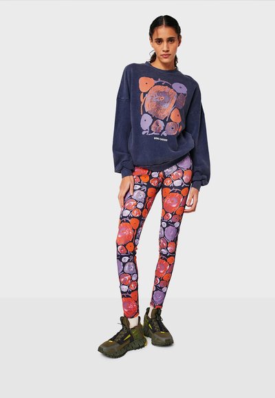 Felpa navy con design grafico; leggings a fantasia vivace in rosso, viola e arancione; sneakers chunky verdi con accenti neri.