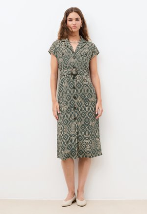 Nainen, joka käyttää vihreä-beigeä kuviollista, napitettavaa mekkoa, jossa on lyhyet hihat ja vyö, seisoo valkoista taustaa vasten.