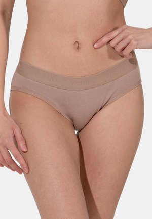 Femme portant des sous-vêtements beiges sans couture avec une large ceinture, main pointant vers sa taille, sur un fond clair uni.