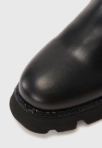 Primo piano della punta di uno stivale di pelle nera con strass neri decorativi lungo il bordo e una robusta suola in gomma nera.