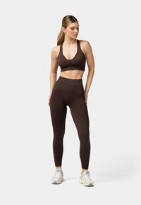 Braune Sport-BH und hochgeschnittene Leggings aus dehnbarem Stoff. Die Modell steht, trägt hellfarbene Sneakers. Neutraler Hintergrund.