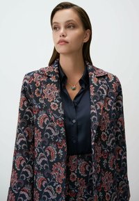 Blazer imprimé en tissu sombre avec des motifs floraux en rouge, rose et bleu. Porté sur une chemise bleu marine à boutons avec un collier à pendentif rond.