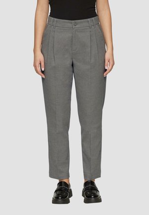 Vrouw die een grijze geplooide broek met hoge taille en zwarte glanzende loafers draagt, staand tegen een effen lichtgrijze achtergrond.