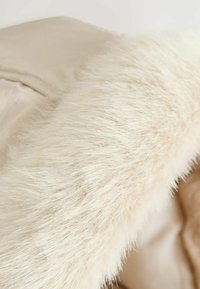 Tessuto esterno beige con un morbido e lungo colletto in pelliccia sintetica bianca. Finitura liscia e tessurizzata sul corpo con cuciture visibili.