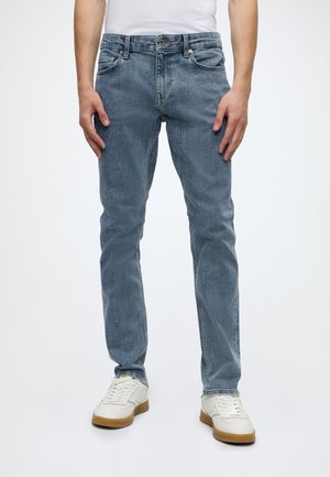 Mann trägt hellblaue Slim-Fit-Jeans, weiße Sneakers mit braunen Sohlen und ein weißes Hemd, steht vor einem weißen Hintergrund.