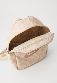 Rucsac bej cu un model abstract piersiciu, din material textil, prevăzut cu două bretele și compartimente cu fermoar. Are logo-ul Nike în nuanță de bej.