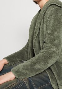 Pälsig, olivgrön, huva-jacka med en mjuk textur; har en lös passform och långa ärmar, kombinerad med mörkblå jeans.