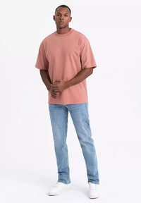 Světle růžové bavlněné tričko s krátkými rukávy, volnější střih, kombinované se světle modrými džíny a bílými teniskami, stojící na jednobarevném pozadí.