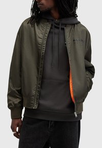 Olivengrøn bomberjakke med en blank overflade, der har lynlås, ribbede manchetter og et orange foer, lagd over en sort hoodie.