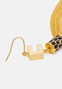 Brinco pendente em tom dourado com um charm em forma de U e um detalhe hexagonal preto. A corrente texturizada adiciona dimensão e interesse visual.