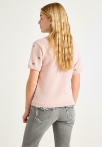 Lyserød, fuzzy kortærmet sweater med broderede sorte sløjfer på skuldrene. Tilsat grå jeans med kontraststing.