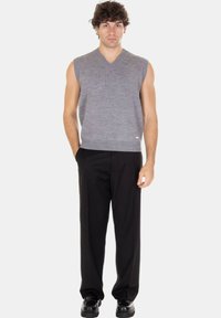 Uomo con capelli ricci che indossa un gilet lavorato a maglia grigio senza maniche, pantaloni neri e scarpe nere, in piedi con una mano in tasca.