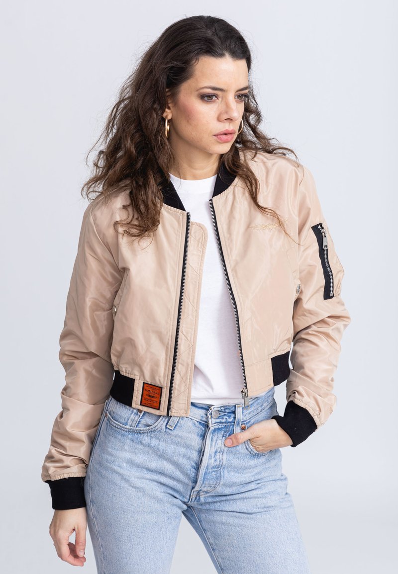 Lichtbeige cropped bomberjack met een ritssluiting aan de voorkant, zwarte ribgebreide manchetten en een oranje label aan de zijkant. Gladde textuur en minimale detailing.