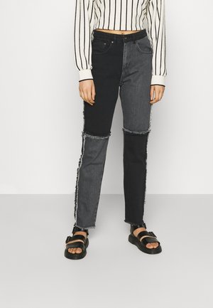 Kvinde iført sorte og mørkegrå patchwork-jeans med flossede kanter, en cropped hvid sweater med sorte striber og sorte sandaler.