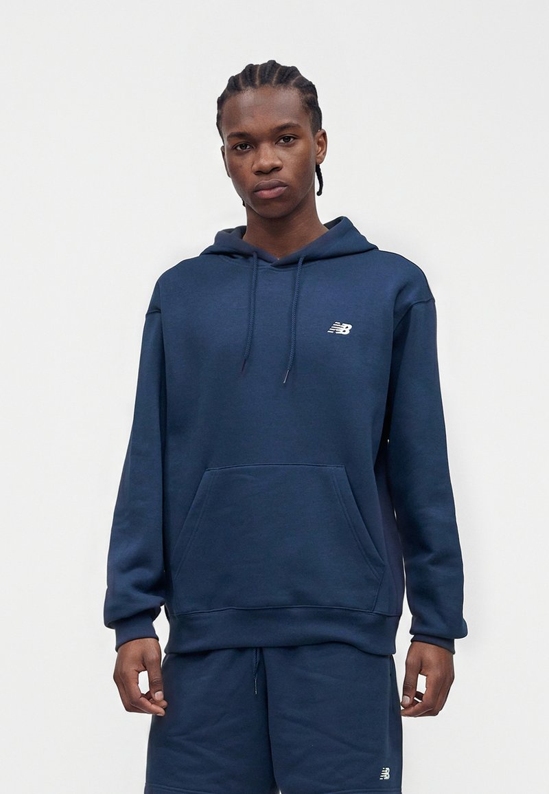 Jong persoon met gevlochten haar, draagbaar een marineblauwe New Balance hoodie en bijpassende korte broek, staand tegen een effen witte achtergrond.