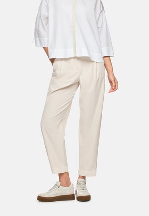 Person wearing loose beige pants, white sneakers with brown soles, and a loose white button-up shirt, standing with one hand in pocket.