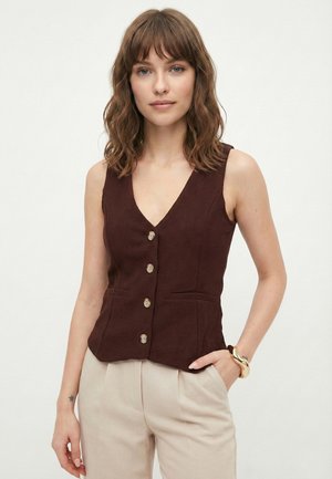 Moteris su banguotais rusvais plaukais dėvi be rankovių rudą liemenę su sagomis ir smėlio spalvos kelnes, dešinėje riešo pusėje – auksinį apyrankę.