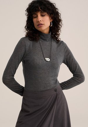 Femme aux cheveux bouclés et foncés portant un col roulé gris, une jupe foncée, un collier en argent et des boucles d'oreilles créoles, debout les mains dans le dos.