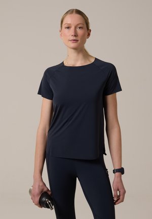 Giovane donna con abbigliamento sportivo blu navy tiene una bottiglia d'acqua, indossa uno smartwatch, in piedi davanti a uno sfondo beige uniforme.