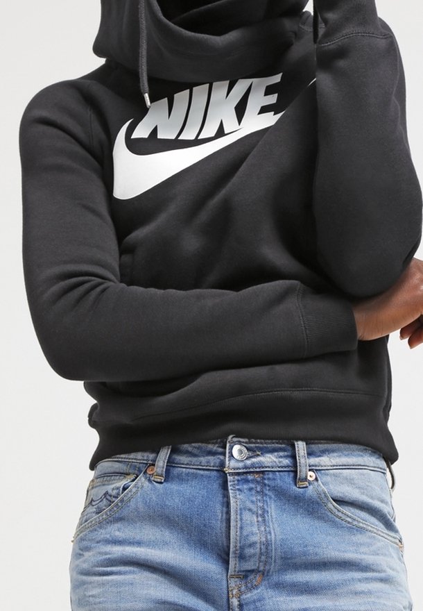 Osoba v černé mikině Nike s bílým logem a světle modrých džínách, se zkříženýma rukama a jednou rukou na tváři.