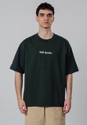 Ung mand med kort krøllet hår iført en mørkegrøn T-shirt med teksten "Still Broke." og beige bukser, stående mod en ensartet lys baggrund.