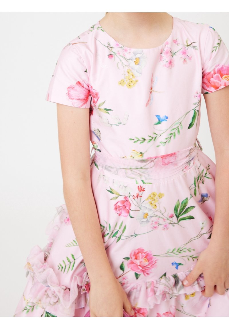 Jeune personne portant une robe fleurie rose avec des manches courtes et des volants, tenant la jupe des deux mains sur un fond uni.