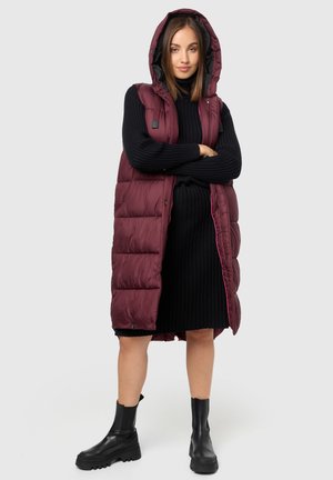 Gilet matelassé bordeaux avec capuche, doté d'une fermeture éclair et de poches latérales, porté sur une robe pull noire à côtes et des bottes noires.