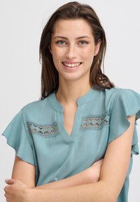Blouse bleu clair en teal avec des manches courtes à volants et un col en V. Présente des détails en dentelle décorative au niveau de la poitrine. Texture de tissu douce.