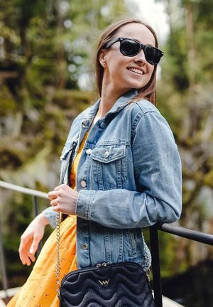Džínová bunda, žluté šaty s květinovým vzorem, černá prošívaná kabelka s řetízkovým popruhem, sluneční brýle a přírodní pozadí.