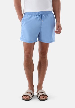 Mann trägt hellblaue Badeshorts mit Kordelzug, weißes Hemd und beige Sandalen, steht vor weißem Hintergrund.