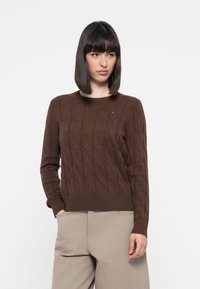 Brown cable-knit sweater with a round neckline, ribbed cuffs, and hem. Small logo detail on the chest, soft texture, fitted silhouette.
