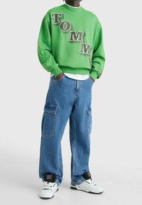 Zelená mikina s bílým a černým nápisem "TOMMY", doplněná světlými modrými volnými cargo džíny a bílými teniskami s černými detaily.