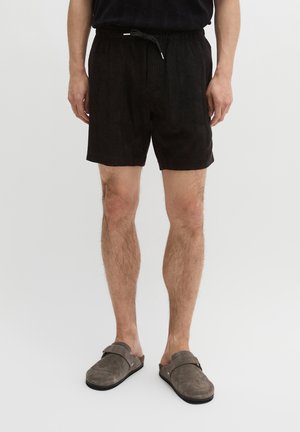 Man die zwarte shorts met koord draagt en grijze instapschoenen, staand op een witte achtergrond.