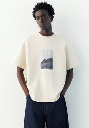 Jeune homme aux cheveux courts et bouclés portant un t-shirt crème oversized avec un motif de maison et un pantalon bleu foncé, debout devant un fond blanc.