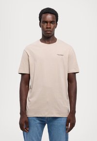T-shirt in cotone beige con maniche corte e scollo rotondo. Presenta un piccolo logo nero sul lato sinistro. Indossato con jeans azzurri chiari.