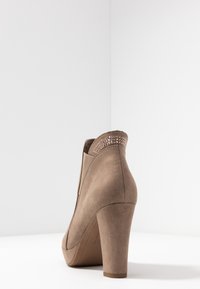 Beige mokkasinivilla nilkkuri, jossa on korkea korko ja koristeellinen niittikiinnitys takana. Sileä pinta ja virtaviivainen muoto.