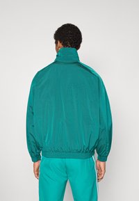 Man vänd bort från kameran, klädd i en teal vindjacka med elastiska muddar och midjeband, matchat med teal träningsbyxor med vita sidoränder.