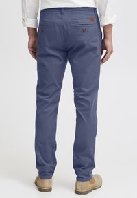 Pantalons chino en tissu bleu foncé avec une texture lisse. Présente des poches arrière avec des boutons marron et un petit patch logo sur la ceinture.